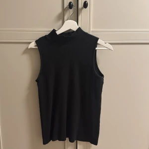 Svart ribbad polotopp från Pieces - Svart ärmlös polotopp från Pieces i ribbad bomullsblandning. Toppen har en hög krage och en slim passform som sitter snyggt mot kroppen. Perfekt att styla med jeans eller kjol för en clean look.