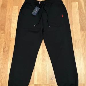 Svarta mjukisbyxor Polo Ralph Lauren - Svarta mjukisbyxor från Polo Ralph Lauren med dragsko i midjan och klassisk röd logga broderad på vänster höft. Byxorna har elastisk midja, ribbade benslut och en bakficka. Perfekta för chill eller streetwear.