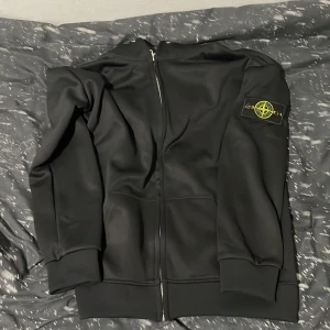 Svart zip hoodie  - Svart hoodie från Stone Island med dragkedja framtill och klassisk logga på vänster ärm. Tröjan har ribbade muddar och är tillverkad i ett mjukt material som känns skönt mot huden. Perfekt för dig som gillar streetwear och vill ha en stilren look.