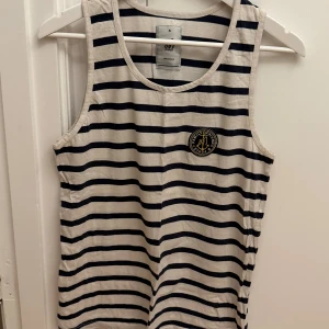 Randig linnetopp från Mouli - Säljer en vit och marinblå randig linnetopp från Mouli i 100% bomull. Toppen är ärmlös med klassisk rund halsringning och har ett broderat märke på bröstet. Perfekt för en chill och stilren look. Storlek S.