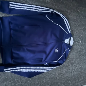 Blå Adidas tröja med vita ränder - Säljer en klassisk marinblå Adidas vindtröja med vita ränder längs ärmarna och vit. Den värmer inte jätte bra men är en riktigt skön tröja generellt. Adidas storlekarna är lite mindre än varligt så även om det står S så skulle jag nog säga att den passar XS lite mer. Priset är på grund av att detta märke inte är så inne längre , men tröjan har ej defekter mm och är i ganska nytt skick.