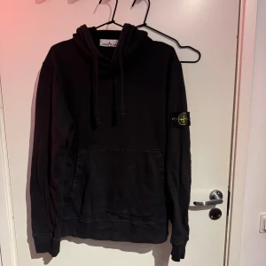 Svart hoodie från Stone Island (KVITTO) - Svart hoodie från Stone Island med klassisk patch på vänster ärm. Tröjan har huva med snörning och en stor magficka framtill. Tillverkad i mjuk bomull, perfekt för chill dagar. Snygg och enkel design som funkar till allt. L men sitter mer som M lite solblekt
