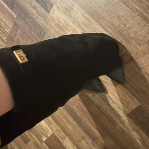 Svarta mockastövlar med spetsig tå - Säljer ett par svarta stövlar i mocka med snygg spetsig tå och hög klack. Stövlarna har en elegant gulddetalj upptill som ger en lyxig känsla. Perfekta för dig som vill sticka ut med en stilren och trendig look.
