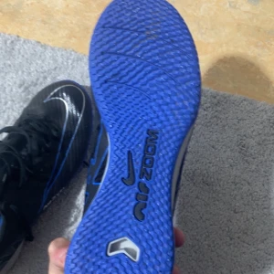 Priset  kan diskuteras  - Säljer ett par Nike Mercurial Zoom fotbollsskor i svart med blå och silverdetaljer. Skorna har snörning, platt sula och är tillverkade i syntetmaterial. Perfekta för inomhusfotboll med sin greppvänliga blå sula och sportig design