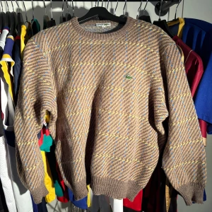 Brun rutig stickad tröja Lacoste - Retro stickad tröja från Lacoste i brunt med rutigt mönster i blått och gult. Klassisk rund halsringning och broderad krokodil på bröstet. Perfekt för dig som gillar vintagevibbar och vill sticka ut med en unik look. Har 3 små hål i ena armen då jag fastnade i en dörr… därav priset 