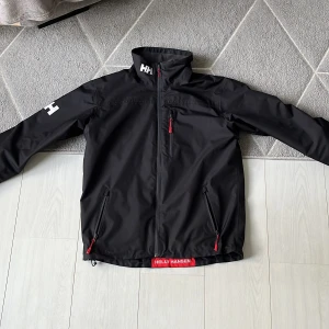 Svart vindjacka från Helly Hansen - Svart vindjacka från Helly Hansen med hög krage, röda dragkedjor och HH-logga på ärm och krage. Inga defekter, förutom att tråden på dragkedjan låsnat (inget som påverkar jackan). Perfekt för blåsiga dagar. Pris kan diskuteras vid snabb affär