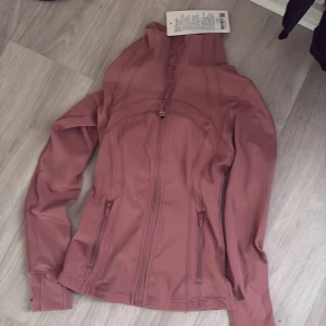 Rosa vindjacka från Lululemon - Snygg rosa vindjacka från Lululemon med figurnära passform. Jackan har dragkedja framtill, två fickor med dragkedja och långa ärmar. Perfekt för träning eller när du vill ha en sportig look. Materialet är mjukt och stretchigt syntet.