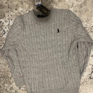 Grå kabelstickad tröja Polo Ralph Lauren - Kabelstickad grå tröja från Polo Ralph Lauren med klassisk rund halsringning och broderad svart logga på bröstet. Tröjan är stickad i mjukt material och har ribbade muddar vid ärmslut och nederkant. Perfekt för lager-på-lager-stil. Helt oanvänd.I bra skick