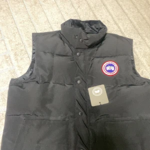 Svart dunväst från Canada Goose - Svart dunväst från Canada Goose med klassisk hög krage och tryckknappar framtill. Västen har två stora fickor och den ikoniska runda loggan på bröstet. Perfekt för lager-på-lager och riktigt snygg till streetwear eller outdoor-stil.