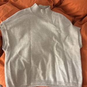 Ljusgrå stickad väst med hög krage - Snygg ljusgrå stickad väst med ribbad hög krage och breda ärmhål. Västen har en loose passform och är perfekt att styla över en t-shirt eller skjorta. Materialet är mjukt och ger en chill vibe till outfiten.