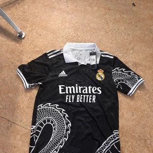 Real Madrid svart draktröja Adidas - Svart Real Madrid fotbollströja från Adidas med vit drakdesign, klubbmärke på bröstet och vit krage. Tröjan har korta ärmar med vita detaljer och klassiska Adidas-ränder på axlarna. Materialet är lätt och andas, perfekt för match eller träning.