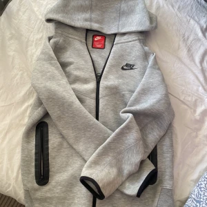 Grå hoodiejacka från Nike med dragkedja - Snygg grå hoodiejacka från Nike med svart logga på bröstet. Jackan har hel dragkedja framtill, två fickor med svarta detaljer och huva. Tillverkad i mjukt material, perfekt för chill eller träning. Svarta muddar vid ärmslut ger en clean look.