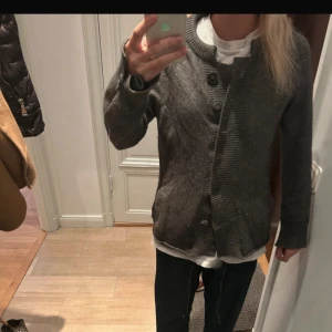 Mörkgrå stickad kofta med knappar - Mysig mörkgrå stickad kofta med grov ribbad kant och stora knappar framtill. Koftan har lång ärm och en rak, lite loose passform. Perfekt att slänga över en t-shirt för en chill look.