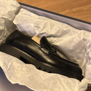 Svarta loafers Vagabond Kenova - Säljer ett par svarta loafers från Vagabond, modell Kenova. Skorna har chunky platt sula med grovt mönster, rund tå och klassisk pennyloafers-detalj. Tillverkade i slätt skinn som ger en stilren och modern look. Perfekta för dig som gillar tidlös design med edge.