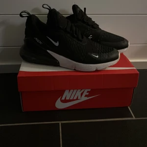 Nike Air Max 270 Svarta Sneakers - Knappt använda svarta Nike Air Max 270 sneakers med vit sula och tydliga Air Max-detaljer. Skorna har snörning, mesh-överdel och en stor synlig luftbubbla i hälen. Snygg och sportig design med vita Nike-loggor på sidorna och tungan. Perfekta för dig som gillar streetstyle. Hela och fräscha skor.