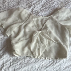 Vit blus med knyt från Lindex ✨✨ - Supersöt vit blus från Lindex med korta puffärmar och omlottknytning framtill. Toppen är croppad och har smockad resår i midjan baktill. Tunt, lätt material som ger en luftig känsla – perfekt för varma dagar. Har en pytteliten lös tråd på baksidan 