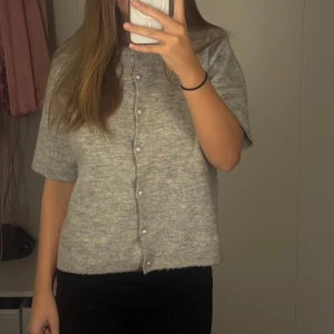 Grå stickad kofta från 157, pärlknappar - Supermysig ljusgrå stickad kofta från 157 med kort ärm och pärlknappar framtill. Koftan har en rak passform och ribbade kanter vid ärmslut och nederkant. Perfekt att slänga över en topp när det är lite kyligt ute.