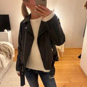 Svart skinnjacka med bälte och dragkedjor - Snygg svart skinnjacka i klassisk bikerstil med silvriga dragkedjor, knappar och bälte i midjan. Jackan har långa ärmar och coola detaljer som axelklaffar och dragkedjor vid ärmsluten. Perfekt för dig som vill ha en edgy look.