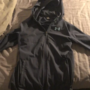 Svart vindjacka från Under Armour - Snygg svart vindjacka från Under Armour med huva och dragkedja framtill. Jackan har logga på bröstet och stort Under Armour-tryck på ryggen. Tillverkad i lätt polyester, perfekt för blåsiga dagar. Fickor med dragkedja och sportig look.