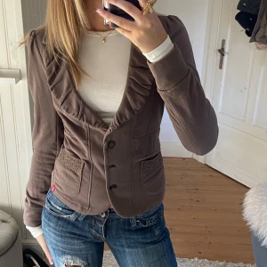 Brun kavaj med volangkrage - Säljer en brun kavaj med snygg volangkrage och dekorativa fickor framtill. Jackan har knäppning med tre knappar och är figursydd för en feminin siluett. Perfekt för dig som vill ha en unik och trendig look. OBS‼️finns en liten fläck på ärmen, skriv privat för bild