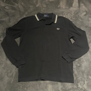 Navy blue långärmad pikétröja Fred Perry - Navy blue långärmad pikétröja från Fred Perry med klassisk vit broderad logga på bröstet och vit rand på kragen. Tröjan är i 100% bomull och har en stilren, tidlös look med knäppning vid halsen. Perfekt för dig som gillar enkel och snygg stil.
