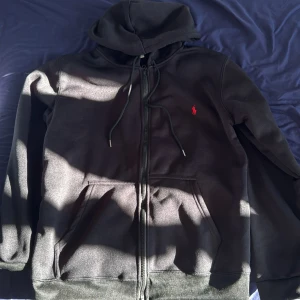 Svart hoodie från Ralph Lauren - Svart hoodie från Ralph Lauren med dragkedja och huva. Klassisk röd logga broderad på bröstet, två fickor framtill och snörning i huvan. Perfekt för chill dagar och enkel att matcha med jeans eller joggers.
