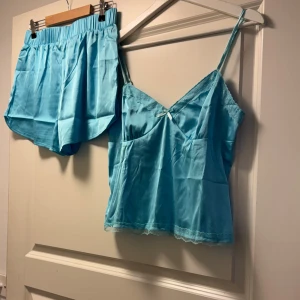 Turkos linnetopp i satin med spets - Säljer en turkos linnetopp i satin med smala axelband och spetsdetaljer vid urringningen och nederkanten. Toppen har en liten rosett framtill och en mjuk, glansig finish som känns lyxig och chill. Perfekt för dig som gillar en feminin och bekväm stil.