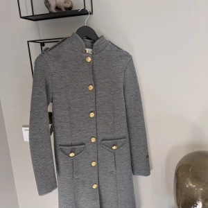 Klassisk Busnelkappa i Grå/mullvad - Skön och mjuk Busnel med raka linjer, ståkrage och dekorativa guldknappar. Toppkvalite!! Kappan har två fickor med lock och ett litet broderat Busnel-märke på ärmen. Klassisk, tidlös design. Strl 36