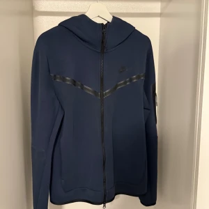 Mörkblå Nike tech fleece - Nike hoodie i mörkblått med hel dragkedja och huva. Snygg detalj med svart V-formad rand över bröstet och diskret Nike-logga. Långärmad modell med ficka på ärmen. Nypris 1200kr. Perfekt för chill eller nu mot hösten/vintern😃 pris kan diskuteras☺️