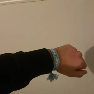 Blått Christian Dior J'Adior armband - Snyggt blått armband från Christian Dior med texten 'J'Adior' broderad i vitt. Armbandet är gjort i tyg och har tofsar i ändarna för en cool och avslappnad look. Perfekt accessoar för att lyfta din outfit med en touch av lyx.