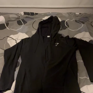 Svart hoodie från Gymshark med dragkedja - Svart hoodie från Gymshark i storlek S. Tröjan har hel dragkedja, huva med snörning och diskret logga på bröstet. Tillverkad i en mix av polyester, bomull och elastan för en skön och flexibel känsla. Perfekt för träning eller chill.