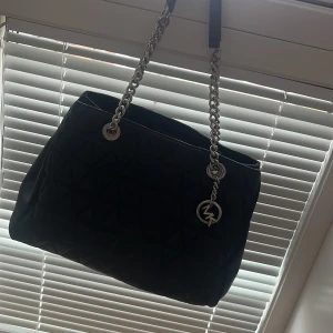 Svart quiltad axelväska Michael Kors - Snygg svart axelväska från Michael Kors i quiltat skinn med silverfärgade kedjehandtag och MK-hänge. Väskan har rymligt fack, innerficka med dragkedja och magnetlås. Perfekt för dig som gillar stilrena detaljer och lyxig känsla.