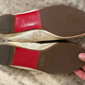 Guldiga ballerina från Louboutin - Snygga ballerina från Christian Louboutin i guldigt skinn med klassisk röd sula och öppen tå. Modellen har rundad form och mjuk insida med Paris-logga. Perfekt för dig som vill ha en lyxig och unik touch på din outfit.