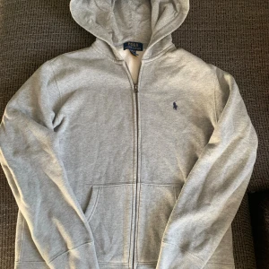 Grå hoodie från Polo Ralph Lauren - Ljusgrå hoodie från Polo Ralph Lauren med dragkedja framtill, huva och ficka. Tröjan har broderad marinblå logga på bröstet och är tillverkad i mjuk bomullsmix. Perfekt för chill dagar och enkel att matcha med jeans eller joggers.