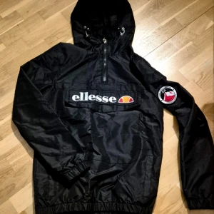 Svart vindjacka från Ellesse - Svart vindjacka från Ellesse med stor logga framtill och patch på ena ärmen. Jackan har huva med snörning, half zip-dragkedja och elastiska muddar. Perfekt för dig som gillar sportig och street-inspirerad stil.