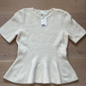 Vit ribbad topp från H&M - Säljer en ljusvit ribbad topp från H&M med korta ärmar och rund hals. Toppen har en peplumformad nederdel som ger en snygg siluett. Materialet är mjukt och stickat, perfekt för att lyfta din look med en clean vibe.