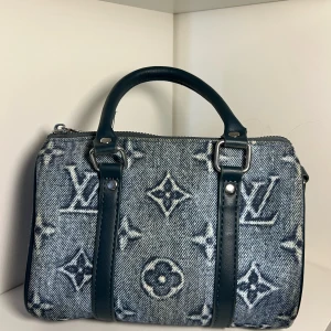 Louis Vuitton denim handväska - Snygg handväska från Louis Vuitton i blått denimtyg med klassiskt monogrammönster. Väskan har svarta skinnhandtag och detaljer i silverfärgad metall. Perfekt storlek för vardagsgrejer och har dragkedja upptill.