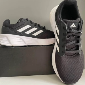 Svarta Adidas sneakers med vit sula - Snygga svarta Adidas sneakers med klassiska vita ränder och vit chunky sula. Ovandelen är i mesh och syntet för bra andningsförmåga. Skorna har snörning och en sportig vibe, perfekta för dig som gillar streetstyle och komfort.