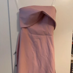 Ljusrosa axelbandslös långklänning - Supersnygg axelbandslös långklänning i ljusrosa med elegant drapering över bysten och en glansig detalj längs sidan. Klänningen har figurnära passform och är tillverkad i ett slätt, satinliknande material som ger en lyxig känsla. Endast använd en gång till bröllop. Skräddarsydd efter någon som är 153-159 cm lång. 