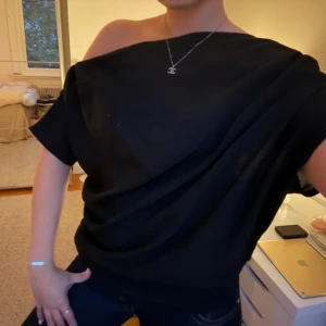 Svart draperad topp från Gina Tricot - Svart draperad topp från Gina Tricot med lös passform och en snygg offshoulder-design. Toppen har korta ärmar och är tillverkad i ett mjukt, lätt material som faller snyggt. Perfekt för dig som gillar en stilren och trendig look.