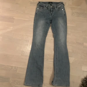 True Religion bootcut jeans blå - Snygga blå bootcut jeans från True Religion, modell Becca Mid Rise Bootcut. Jeansen har klassiska kontrastsömmar, coola fickdetaljer med knappar bak och en tvättad look. Tillverkade i jeansmaterial med medelhög midja.