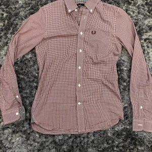 Fred perry skjorta  - Snygg rutig skjorta från Fred Perry i vinrött och vitt. Klassisk button-down krage, långärmad och med knappar framtill. Broderad logga på bröstfickan. Perfekt för dig som gillar stilrena och tidlösa plagg.