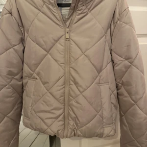 Beige quiltad pufferjacka  -  beige pufferjacka  med dragkedja framtill och två fickor. Jackan har lång ärm och en clean, stilren look som passar perfekt till kyliga dagar. Snyggt rutmönster och lätt oversized passform. Storlek 170 men passar mig som är 164 lång och bär vanligtvis xs-s,Priset går att diskutera om