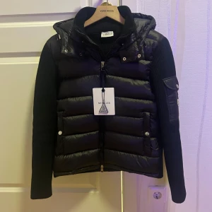 Moncler Cardigan  - Säljer en svart dunväst från Moncler med stickade ärmar och avtagbar huva. Västen har dragkedja framtill, fickor med tryckknappar och en snygg Moncler-logga på ärmen. Perfekt för lager-på-lager och riktigt stilren look.