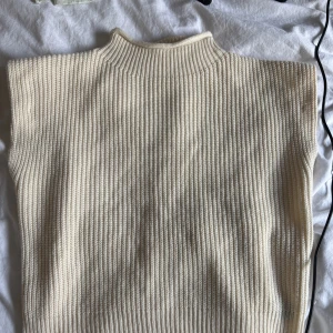 Beige ribbad stickad väst - En stilren beige stickad väst med ribbad struktur och hög krage. Västen är ärmlös och har en rak, något boxig passform som ger en modern look. Perfekt att slänga över en skjorta eller t-shirt för en trendig layering-effekt.