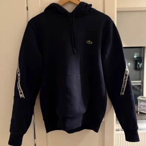 Mörkblå hoodie från Lacoste - Säljer en mörkblå hoodie från Lacoste med klassisk passform. Tröjan har huva med snörning, stor magficka och vita Lacoste-loggor längs ärmarna. Perfekt för dig som gillar stilren streetwear med sportig vibe.