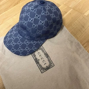Blå keps från Gucci med GG-mönster - Säljer en blå keps från Gucci i storlek M, täckt av det klassiska GG-monogrammet. Kepsen har böjd skärm och justerbar rem bak. Materialet är kraftig bomullscanvas med exklusiv känsla. Perfekt för dig som vill sticka ut med en ikonisk accessoar.