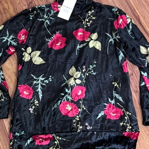 Svart blommig sammetströja Dowry M - Svart långärmad tröja från Dowry i mjuk sammet med stora röda blommor och gröna blad. Tröjan har en half zip-dragkedja framtill och hög krage. Perfekt för dig som gillar unika och färgstarka plagg med skön känsla.