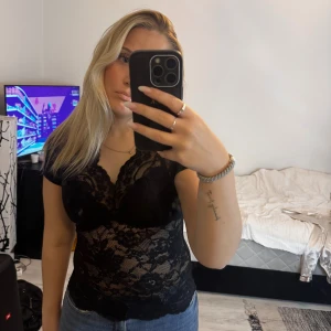 Svart spetsblus med v-ringning - Säljer en svart, genombruten spetsblus med v-ringning och korta ärmar. Blusen har ett blommigt spetsmönster och sitter tight mot kroppen. Perfekt att styla med jeans eller kjol för en trendig look.