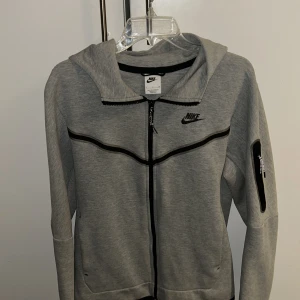 Grå Nike hoodie med dragkedja - Snygg grå hoodie från Nike med svart logga på bröstet och svarta detaljer. Tröjan har hel dragkedja framtill, huva och en praktisk ficka med dragkedja på ärmen. Perfekt för en sportig och avslappnad stil.